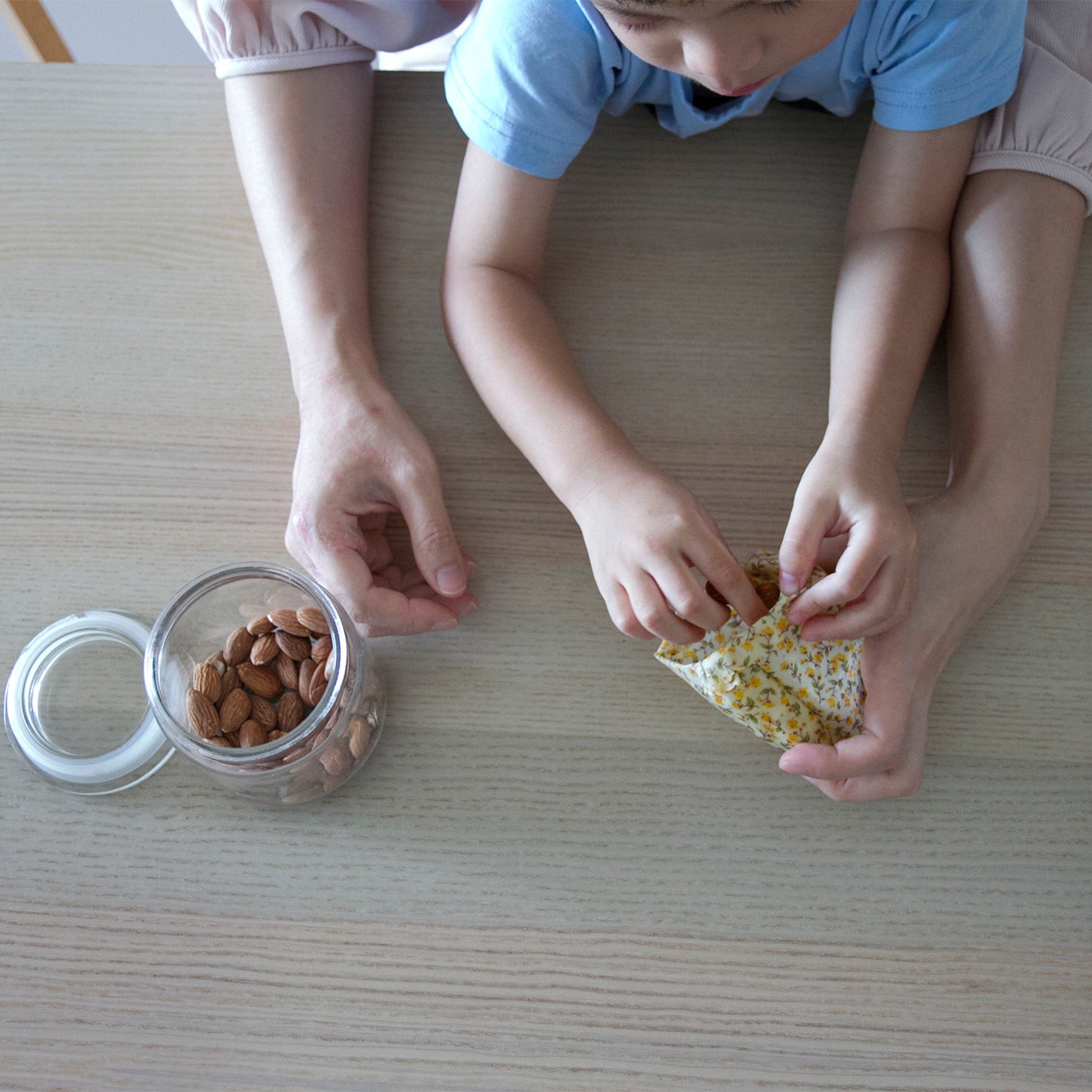 Two hands, one mother and one child, storing almonds in a beeswax-wrap-made-pouch on a wooden surface.